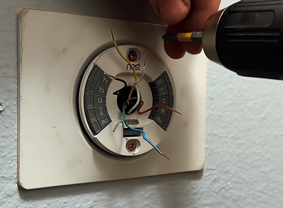 Round smart-thermostat base on a rectangular wall plate, with terminal blocks, loose colored wires, two mounting screws, and a drill nearby.